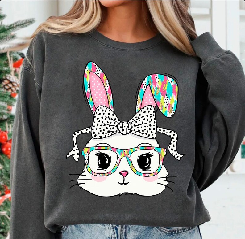May include: A gray sweatshirt with a colorful cartoon bunny wearing glasses and a bow. The bunny has pink ears with white polka dots, a white bow with black polka dots, and black-rimmed glasses with colorful floral accents.