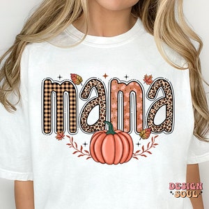 May include: White t-shirt with the word "mama" in a fall-themed design. The letters are decorated with plaid, leopard print, and floral patterns. A pumpkin with fall leaves is below the word.