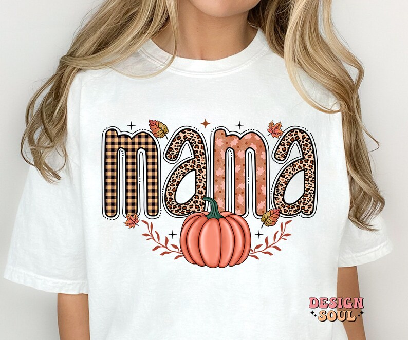 May include: White t-shirt with the word "mama" in a fall-themed design. The letters are decorated with plaid, leopard print, and floral patterns. A pumpkin with fall leaves is below the word.