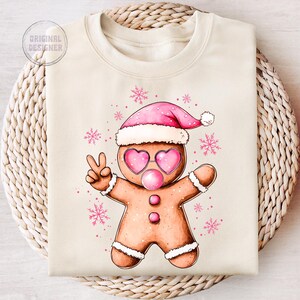 May include: A beige sweatshirt with a gingerbread man wearing pink sunglasses and a pink Santa hat. The gingerbread man is holding up two fingers in a peace sign. The background is white with pink snowflakes.