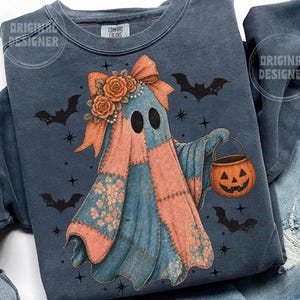 May include: A blue sweatshirt featuring a patchwork ghost design with a pumpkin bucket. The ghost has a bow, floral accents, and black eyes. Bats and stars are scattered around the ghost, creating a Halloween theme.