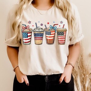 May include: A white t-shirt with a graphic of four iced coffee cups decorated with the American flag. The cups are arranged in a row and have red, white, and blue stripes.