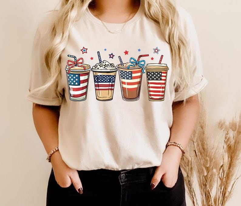 May include: A white t-shirt with a graphic of four iced coffee cups decorated with the American flag. The cups are arranged in a row and have red, white, and blue stripes.