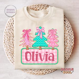 May include: Cream-colored sweatshirt featuring three Christmas trees with pink bows and a teal polka-dot tree. The name "Olivia" is displayed on a pink and white checkered background. The design includes gold glitter accents.