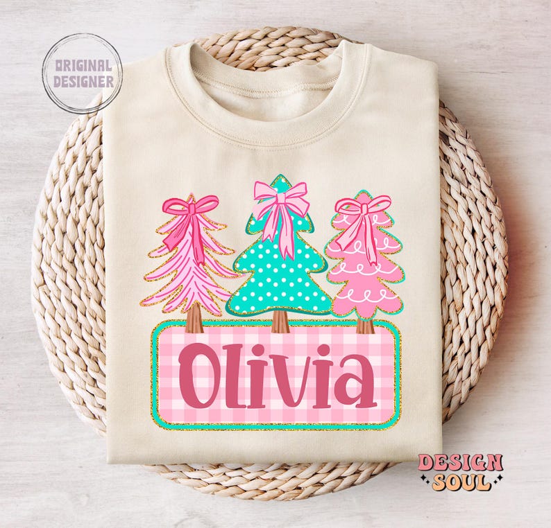 May include: Cream-colored sweatshirt featuring three Christmas trees with pink bows and a teal polka-dot tree. The name "Olivia" is displayed on a pink and white checkered background. The design includes gold glitter accents.