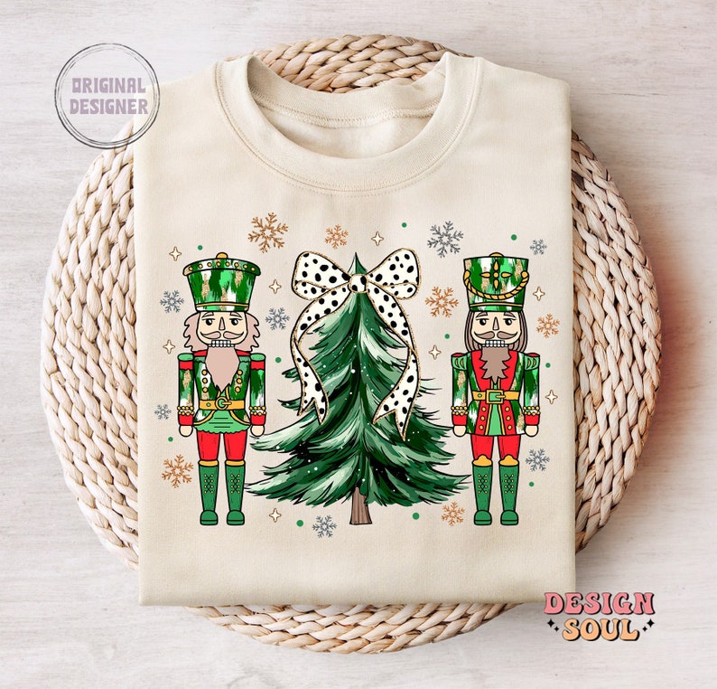 May include: Cream-coloured sweatshirt featuring a Christmas tree with a polka-dot bow, flanked by two nutcracker figures in green and red uniforms. The design includes snowflakes and the text "DESIGN SOUL".