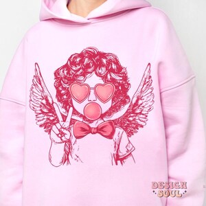 May include: Pink hoodie featuring a graphic of a cherub with angel wings, wearing heart-shaped sunglasses and blowing a bubble. The cherub is wearing a bow tie. The hoodie has the text "DESIGN SOUL" printed on the bottom.