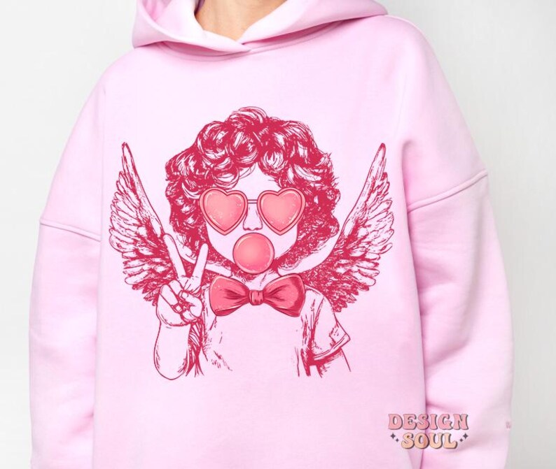 May include: Pink hoodie featuring a graphic of a cherub with angel wings, wearing heart-shaped sunglasses and blowing a bubble. The cherub is wearing a bow tie. The hoodie has the text "DESIGN SOUL" printed on the bottom.