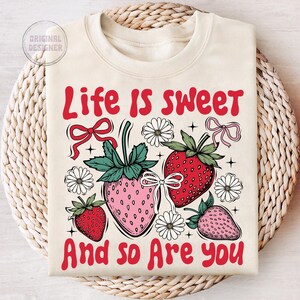 May include: A cream-colored sweatshirt with the text "Life is sweet and so are you" in red. The design features strawberries, flowers, and bows.