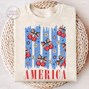 May include: A cream-colored sweatshirt with a blue and white striped design featuring red cherries and stars. The word "AMERICA" is printed in red at the bottom of the shirt.