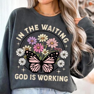 May include: A gray sweatshirt with a graphic print of a butterfly and flowers. The text "In the waiting, God is working" is printed on the sweatshirt.
