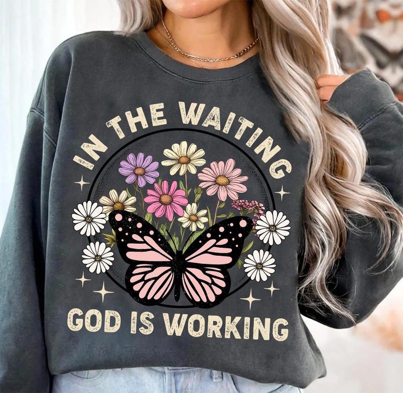 May include: A gray sweatshirt with a graphic print of a butterfly and flowers. The text "In the waiting, God is working" is printed on the sweatshirt.