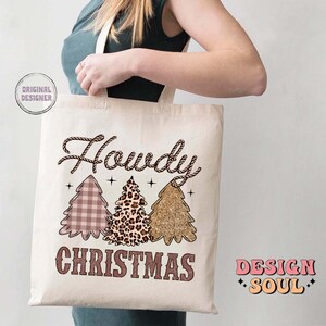 May include: A white canvas tote bag with a brown rope design and the text "Howdy Christmas" in brown lettering. The bag features three Christmas trees with different patterns: plaid, leopard print, and glitter. The text "Design Soul" is printed in pink and orange on the right side of the bag.