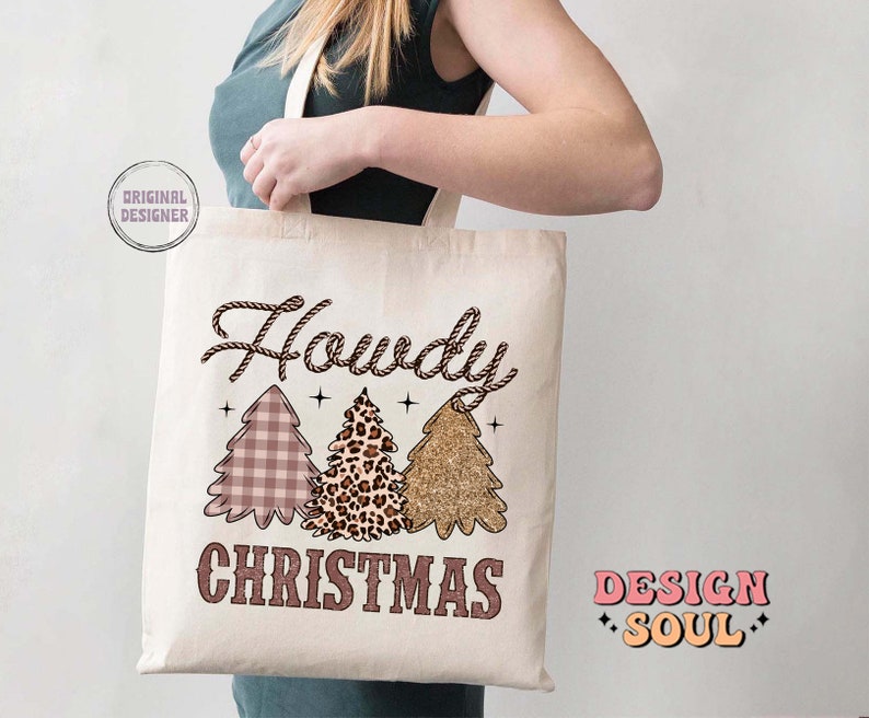 May include: A white canvas tote bag with a brown rope design and the text "Howdy Christmas" in brown lettering. The bag features three Christmas trees with different patterns: plaid, leopard print, and glitter. The text "Design Soul" is printed in pink and orange on the right side of the bag.