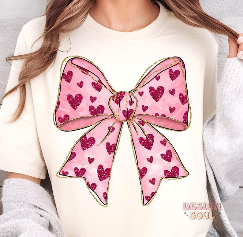 May include: A pink bow with gold glitter accents and a pattern of pink hearts. The bow is on a cream-colored t-shirt.
