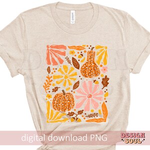 May include: A beige t-shirt with a digital print of a floral design featuring pumpkins and gourds in shades of orange, yellow, pink, and brown. The design is surrounded by leaves and vines.