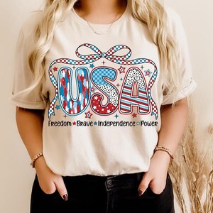 Peut inclure: Un t-shirt blanc avec un graphique USA rouge, blanc et bleu. Le graphique pr&eacute;sente un n&oelig;ud et des &eacute;toiles. Le texte "Freedom Brave Independence Power" est imprim&eacute; sous le graphique.