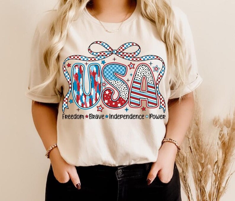 Peut inclure: Un t-shirt blanc avec un graphique USA rouge, blanc et bleu. Le graphique pr&eacute;sente un n&oelig;ud et des &eacute;toiles. Le texte "Freedom Brave Independence Power" est imprim&eacute; sous le graphique.