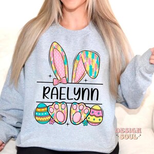 May include: A light gray sweatshirt with a colorful Easter bunny design. The bunny ears are pink, yellow, and blue with white polka dots. The bunny has paw prints and the name "Raelynn" is printed below the bunny.