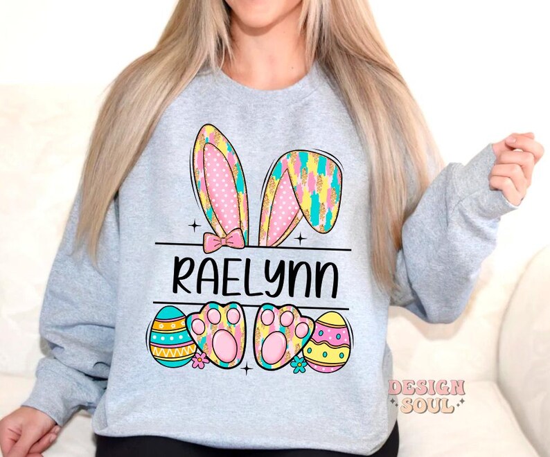 May include: A light gray sweatshirt with a colorful Easter bunny design. The bunny ears are pink, yellow, and blue with white polka dots. The bunny has paw prints and the name "Raelynn" is printed below the bunny.