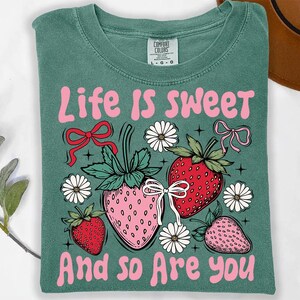 May include: A teal green t-shirt with the text "Life is Sweet and so are you" in pink lettering. The design features three pink and red strawberries with green leaves and white flowers.