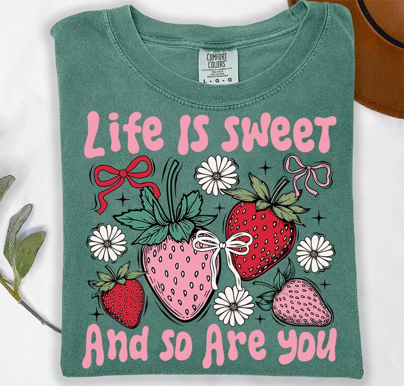 May include: A teal green t-shirt with the text "Life is Sweet and so are you" in pink lettering. The design features three pink and red strawberries with green leaves and white flowers.