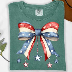 May include: A teal blue t-shirt with a large red, white, and blue bow with stars printed on it. The bow has a string of beads hanging from it with stars at the end of each string.