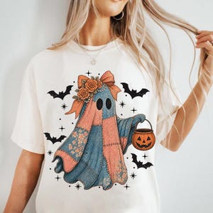 May include: Cream-colored t-shirt featuring a whimsical ghost design. The ghost is adorned with a patchwork quilt-like pattern in shades of blue and peach, a floral bow, and a jack-o'-lantern bucket. Black bats and stars complete the Halloween theme.