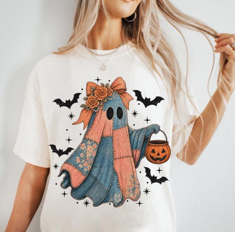 May include: Cream-colored t-shirt featuring a whimsical ghost design. The ghost is adorned with a patchwork quilt-like pattern in shades of blue and peach, a floral bow, and a jack-o'-lantern bucket. Black bats and stars complete the Halloween theme.