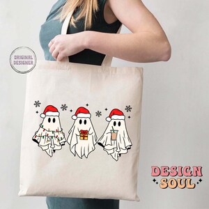 May include: A white tote bag with a design of three ghosts wearing Santa hats. The ghosts are holding a gift, a cup of coffee, and a string of lights. The text "Design Soul" is printed on the bag.