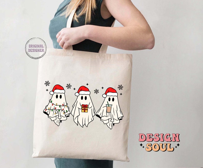 May include: A white tote bag with a design of three ghosts wearing Santa hats. The ghosts are holding a gift, a cup of coffee, and a string of lights. The text "Design Soul" is printed on the bag.