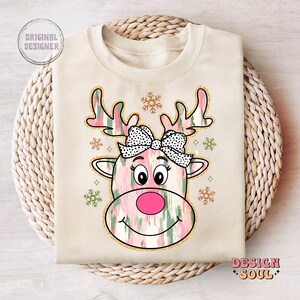 May include: A beige sweatshirt with a pink and green reindeer design. The reindeer has a black and white polka dot bow and a pink nose. The design is surrounded by gold glitter snowflakes.