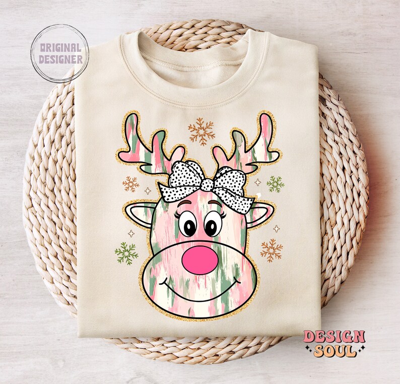 May include: A beige sweatshirt with a pink and green reindeer design. The reindeer has a black and white polka dot bow and a pink nose. The design is surrounded by gold glitter snowflakes.