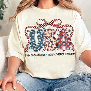 May include: A white t-shirt with a red, white, and blue USA graphic. The graphic features a bow with checkered pattern and the letters are decorated with red, white, and blue flowers. The text "Freedom Brave Independence Power" is printed below the graphic.