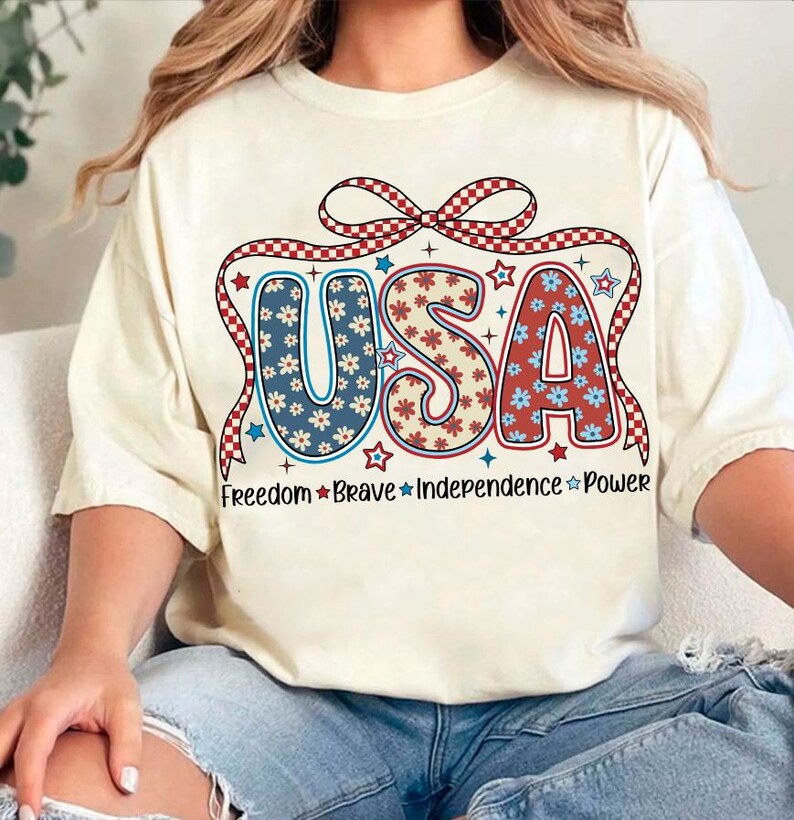 May include: A white t-shirt with a red, white, and blue USA graphic. The graphic features a bow with checkered pattern and the letters are decorated with red, white, and blue flowers. The text "Freedom Brave Independence Power" is printed below the graphic.