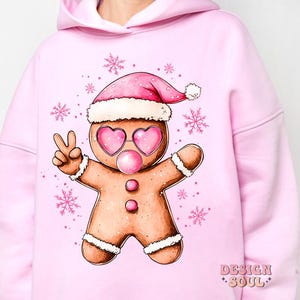 May include: Pink hoodie with a gingerbread man wearing a pink Santa hat and sunglasses, blowing a bubble. The gingerbread man is holding up two fingers in a peace sign. The hoodie has the text "Design Soul" on the bottom right corner.