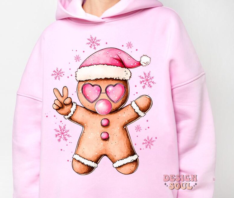 May include: Pink hoodie with a gingerbread man wearing a pink Santa hat and sunglasses, blowing a bubble. The gingerbread man is holding up two fingers in a peace sign. The hoodie has the text "Design Soul" on the bottom right corner.