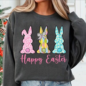 May include: A gray sweatshirt with a pink, blue, and yellow bunny rabbit design. The bunnies are wearing bows and have different patterns. The text "Happy Easter" is printed in pink below the bunnies.