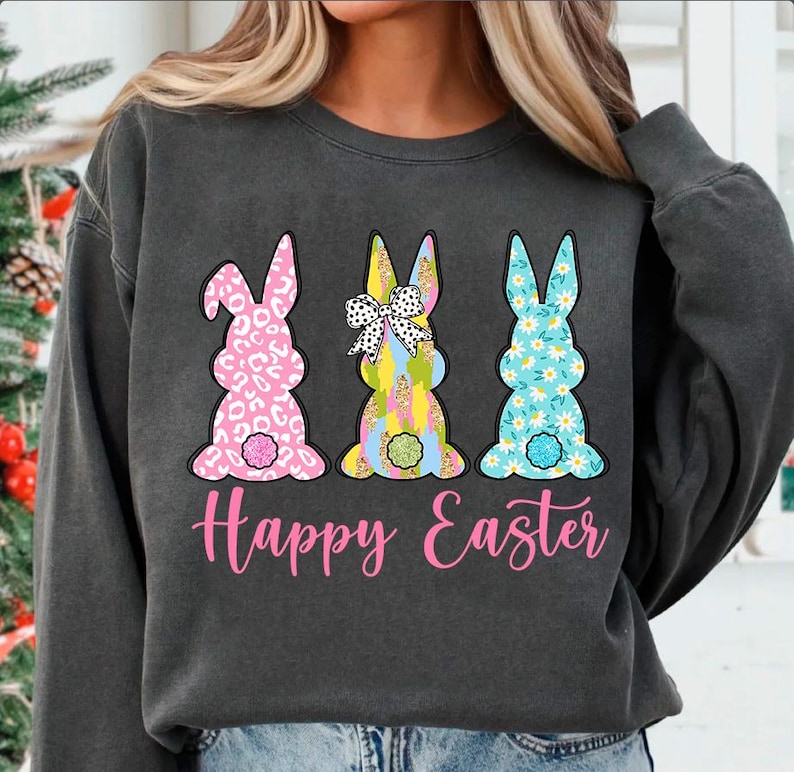 May include: A gray sweatshirt with a pink, blue, and yellow bunny rabbit design. The bunnies are wearing bows and have different patterns. The text "Happy Easter" is printed in pink below the bunnies.