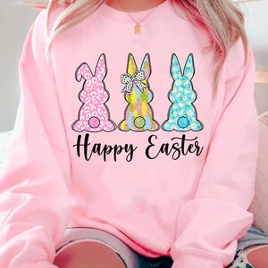 May include: Pink sweatshirt with a graphic of three Easter bunnies with different patterns. The bunnies are facing away from the viewer and the text "Happy Easter" is printed below them.