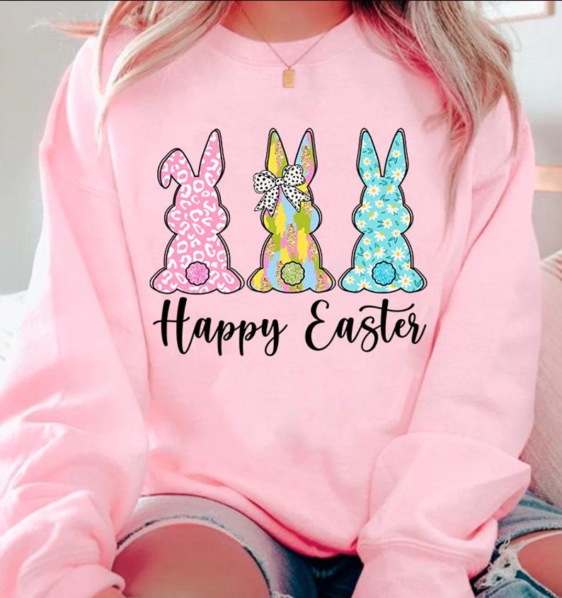 May include: Pink sweatshirt with a graphic of three Easter bunnies with different patterns. The bunnies are facing away from the viewer and the text "Happy Easter" is printed below them.