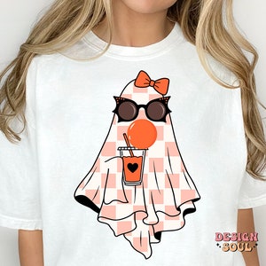 May include: White t-shirt with a pink and white checkered ghost wearing sunglasses and a pink bow. The ghost is holding a pink and orange drink with a straw.
