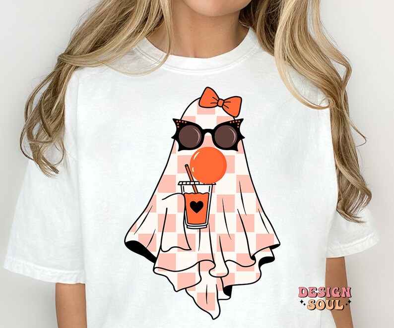 May include: White t-shirt with a pink and white checkered ghost wearing sunglasses and a pink bow. The ghost is holding a pink and orange drink with a straw.
