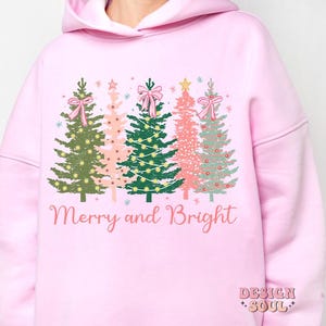 May include: Pink hoodie with a graphic of four Christmas trees with pink bows and the text "Merry and Bright".