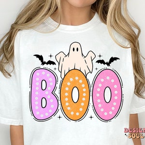 Op de afbeelding: Wit T-shirt met een roze, oranje en witte "BOO"-afbeelding. De afbeelding toont een geest, vleermuizen en stippen.