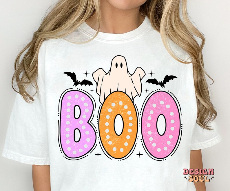 Op de afbeelding: Wit T-shirt met een roze, oranje en witte "BOO"-afbeelding. De afbeelding toont een geest, vleermuizen en stippen.