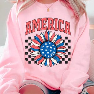 May include: Pink sweatshirt with a distressed graphic of a sunflower with red, white, and blue colors, resembling the American flag. The word "AMERICA" is printed above the sunflower.