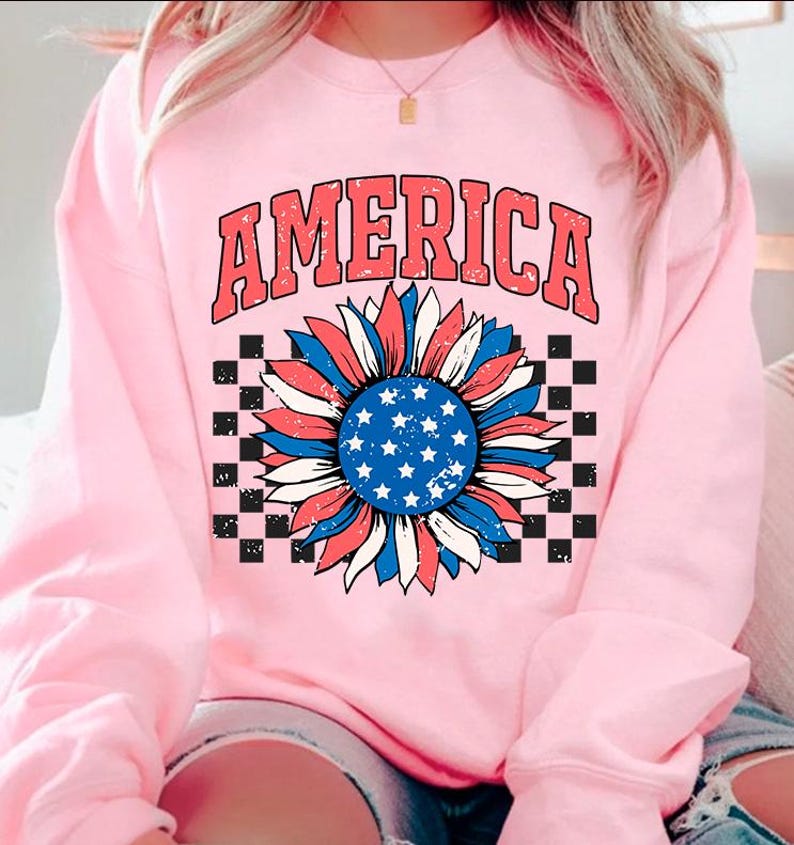 May include: Pink sweatshirt with a distressed graphic of a sunflower with red, white, and blue colors, resembling the American flag. The word "AMERICA" is printed above the sunflower.