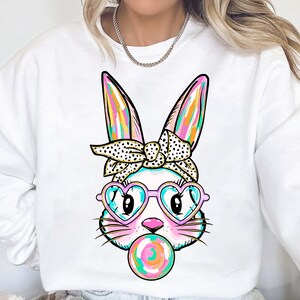 May include: White sweatshirt featuring a colourful cartoon bunny wearing heart-shaped sunglasses and a polka dot headband. The bunny is holding a pink and blue lollipop.