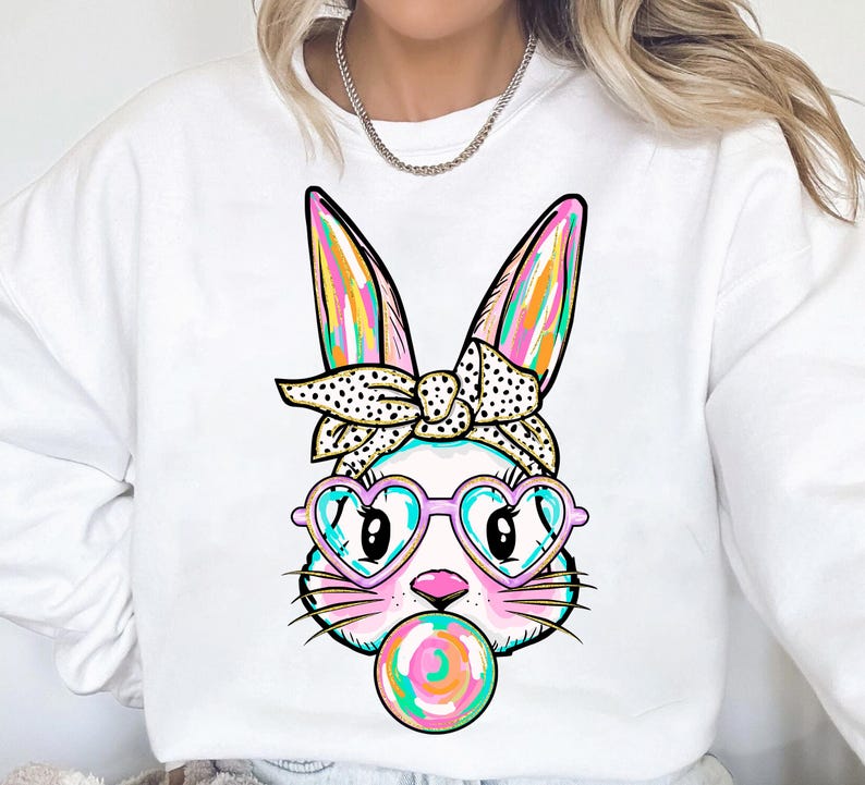 May include: White sweatshirt featuring a colourful cartoon bunny wearing heart-shaped sunglasses and a polka dot headband. The bunny is holding a pink and blue lollipop.